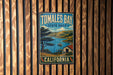 A vintage-style metal sign for Tomales Bay State Park in California, featuring a scenic landscape with a lake, mountains, and a small boat.