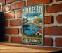 A vintage-style metal sign for Tomales Bay State Park in California, featuring a scenic landscape with a lake, mountains, and a small boat.