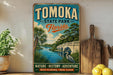 A vintage-style metal sign for Tomoka State Park, Florida, featuring a scenic illustration of a river, palm trees, and a bridge.