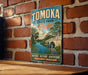 A vintage-style metal sign for Tomoka State Park in Florida, featuring a scenic illustration of a bridge, palm trees, and a bird, mounted on a brick wall.