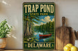 A vintage metal sign for Trap Pond State Park in Delaware, featuring a painting of a red canoe on a pond surrounded by trees and a cloudy sky.