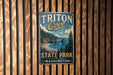 A vintage metal sign for Triton Cove State Park in Washington, featuring a scenic landscape with pine trees, a beach, and a body of water.