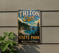 A vintage metal sign for Triton Cove State Park in Washington, featuring a scenic landscape with a beach, mountains, and pine trees.