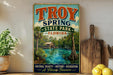 A vintage-style sign for Troy Spring State Park in Florida, featuring a scenic image of a river, palm trees, and a wooden cabin.