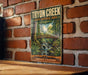 A vintage-style metal sign for the "Tryon Creek State Natural Area" in Oregon, featuring a scenic illustration of a bridge, trees, and a stream.