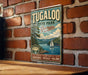 A vintage-style metal sign for Tugaloo State Park in Georgia, featuring a scenic landscape with a lake, mountains, and a sailboat.