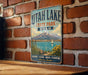 A vintage-style metal sign for Utah Lake State Park, featuring a scenic mountain landscape with a lake and a sailboat.