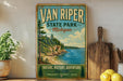 A vintage-style sign for Van Riper State Park in Michigan, featuring a scenic landscape with a lake and mountains.