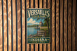 A vintage metal sign for Versailles State Park, featuring a scenic illustration of a lake, boats, and a lighthouse, is displayed on a wooden wall.