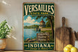 A vintage-style metal sign for Versailles State Park in Indiana, featuring a scenic image of a lake and a lighthouse.