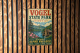 A vintage metal sign for Vogel State Park in Georgia, featuring a scenic illustration of a mountain landscape with a cabin and flowers.