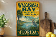 A vintage-style metal sign for Waccasass Bay Preserve, Florida, featuring a bird and palm trees, is displayed on a shelf next to a wooden cutting board and some lemons.