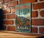 A vintage-style metal sign for Waimea Canyon State Park in Kauai, Hawaii, featuring a scenic mountain landscape.