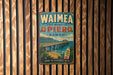 A vintage-style metal sign for Waimea Pier in Hawaii, featuring a wooden pier and a surfboard, is displayed on a wooden wall.