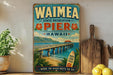 A vintage-style poster of Waimea Pier in Hawaii, featuring a wooden pier, palm trees, and a surfboard, is displayed on a wall next to a wooden cutting board and some lemons.