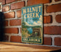 A vintage-style metal sign for Walnut Creek State Park in Oklahoma, featuring a scenic landscape with a lake and mountains.