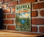 A vintage metal sign for Warwick State Forest in Massachusetts, featuring a scenic landscape with pine trees, a lake, and mountains.