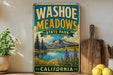 A vintage-style metal sign for Washoe Meadows State Park in California, featuring a scenic landscape with mountains, a lake, and wildflowers.