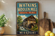 A vintage-style sign for Watkins Woolen Mill State Park in Missouri, featuring an illustration of a mill with a waterfall.