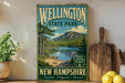 A vintage-style metal sign for Wellington State Park in New Hampshire, featuring a scenic landscape with a lake, mountains, and pine trees.