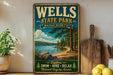 A vintage-style metal sign for Wells State Park in Massachusetts, featuring a scenic landscape with a lake, pine trees, and a beach.