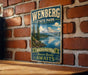 A vintage-style metal sign for Wenberg State Park, featuring a scenic mountain landscape with a lake and pine trees.