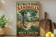 A vintage-style metal sign advertising Weymouth Woods and Sandhills, featuring a scenic illustration of a forest and a bird perched on a tree.