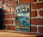 A vintage-style metal sign for Whitefish Lake State Park in Montana, featuring a scenic landscape with a lake, mountains, and a boat on the water.