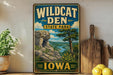 A vintage-style sign for Wildcat Den State Park in Iowa, featuring a scenic landscape with a river, mountains, and trees.