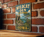 A vintage-style metal sign for Wildcat Den State Park in Iowa, featuring a scenic landscape with a river and mountains.