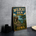 A vintage-style metal sign for Wildcat Den State Park in Iowa, featuring a scenic landscape with a rocky cliff and a river.