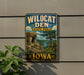 A vintage-style metal sign for Wildcat Den State Park in Iowa, featuring a scenic landscape with a rocky cliff, trees, and a river.