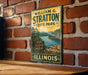 A vintage-style metal sign for William G. Stratton State Park in Illinois, featuring a scenic landscape with a river and mountains.