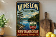 A vintage-style sign for Winslow State Park in New Hampshire, featuring a scenic landscape with pine trees, a lake, and mountains.