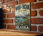 A vintage-style metal sign for Wood-Tikik State Park in Alaska, featuring a scenic landscape with a plane and mountains in the background.