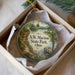 A circular ornament with a scenic landscape featuring a river, a bridge, and a forest, hanging from a string.