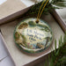 A decorative ornament in a box, featuring a scenic landscape with a lake, mountains, and a wooden dock.