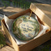 A decorative ornament in a wooden box, featuring a scenic landscape with a river, trees, and a bridge.