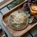 A wooden tray with a decorative ornament featuring a scenic landscape of Abilene State Park, Texas, and pine cones.