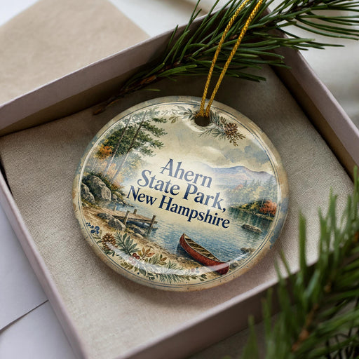 A decorative ornament in a box, featuring a scenic landscape with a lake, mountains, and a canoe, with the text "Ahern State Park, New Hampshire" printed on it.