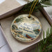 A decorative ornament in a box, featuring a scenic landscape with a lake, mountains, and a canoe, with the text "Ahern State Park, New Hampshire" printed on it.