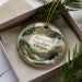 A decorative ornament in a box, featuring a scenic landscape with mountains, trees, and a waterfall.