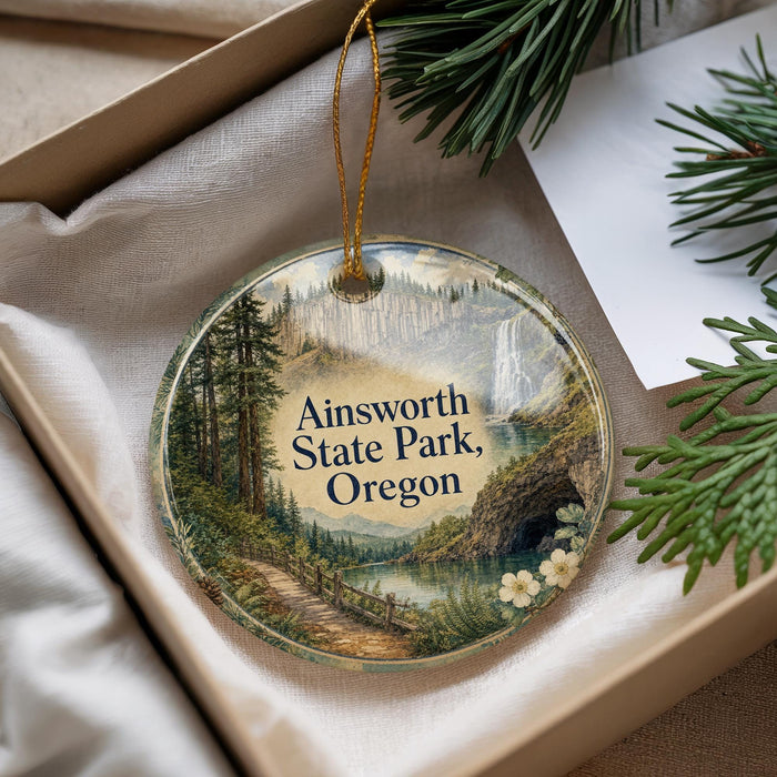 A decorative ornament with a scenic landscape of Ainsworth State Park, Oregon, featuring a winding path, a waterfall, and pine trees.