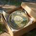 A round ornament in a box, featuring a scenic landscape with mountains, trees, and a winding path.