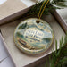 A decorative ornament in a box, featuring a scenic landscape with mountains, a lake, and pine trees.