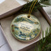 A decorative ornament in a box, featuring a landscape scene with pine trees, flowers, and a bird.