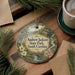 A decorative ornament with a scenic landscape of Andrew Jackson State Park, South Carolina, featuring a bridge, water, and flowers. The ornament is hanging on a wooden surface, surrounded by pine branches and a cup of coffee.
