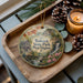 A decorative ornament featuring a scenic landscape of Babcock State Park, West Virginia, with a waterfall and pine trees. The ornament is placed on a wooden tray, accompanied by pine cones and a lit candle.