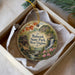 A decorative ornament in a box, featuring a scenic landscape with a waterfall, flowers, and a house.