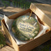 A round ornament in a brown box, featuring a scenic landscape with a bird and flowers, is displayed on a wooden surface.